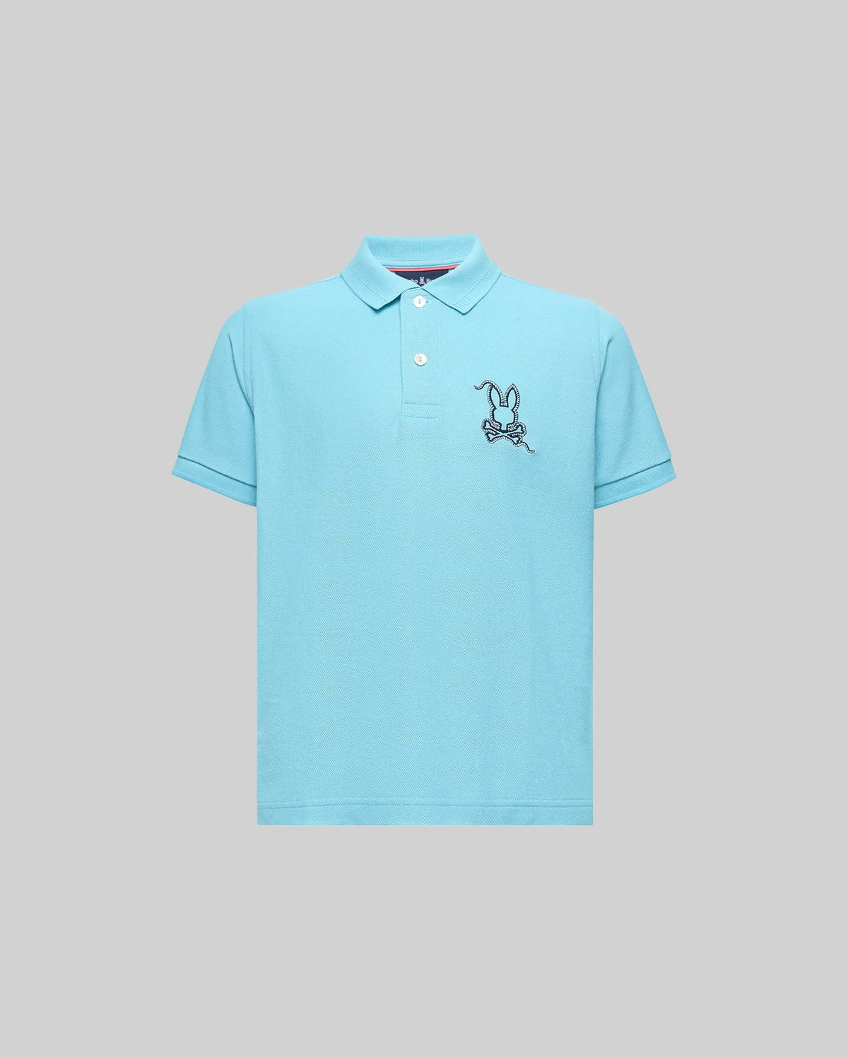 The KIDS CYRUS PIQUE POLO (KT0100031) by Psycho Bunny is a light blue short-sleeve Pima cotton polo with a two-button placket and an embroidered cartoon bunny on a skateboard, shown on a plain gray background.
