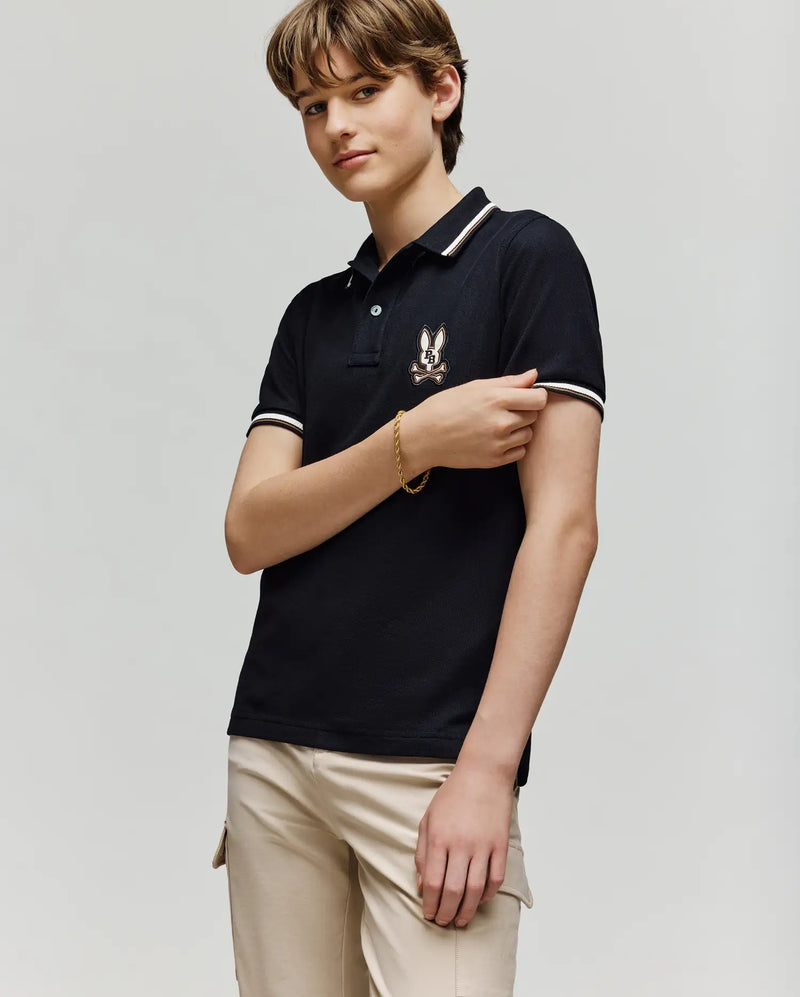 A young person with short, light brown hair wears the Psycho Bunny KIDS WADE PIQUE POLO - KT0100032, featuring an embroidered logo on the chest, paired with beige pants and set against a plain light gray background.