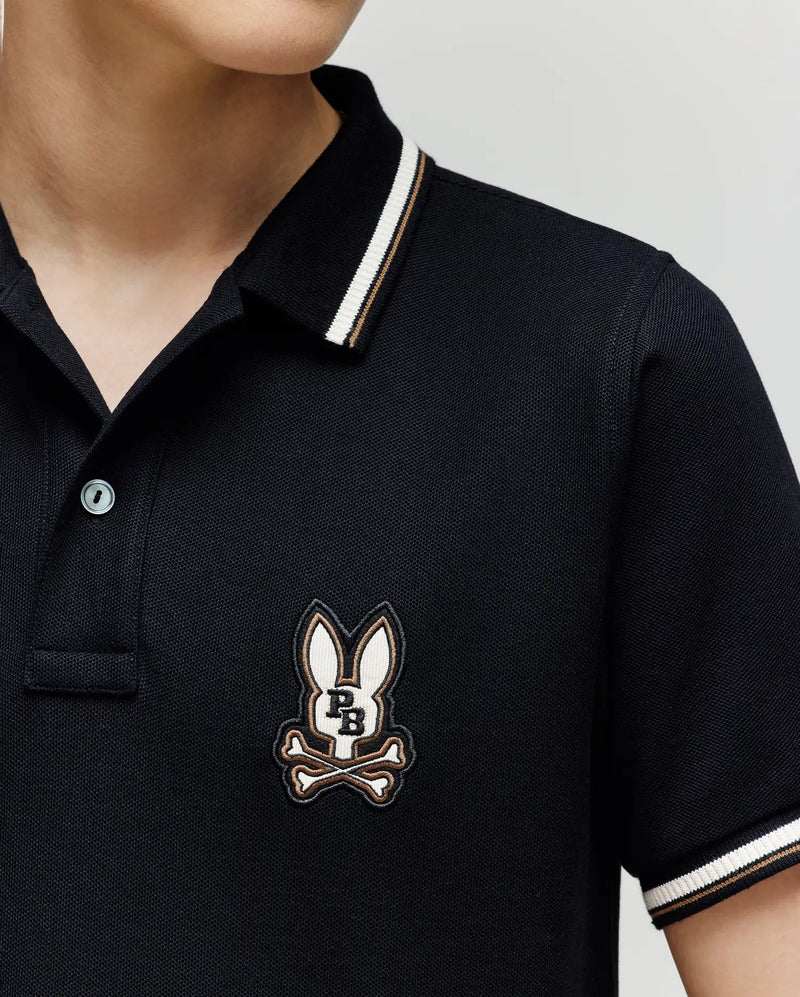 A child wears the Psycho Bunny KIDS WADE PIQUE POLO (KT0100032), a black Pima cotton polo with white colorblock tipping on the collar and cuffs, featuring an embroidered bunny head above crossed bones on the left chest.