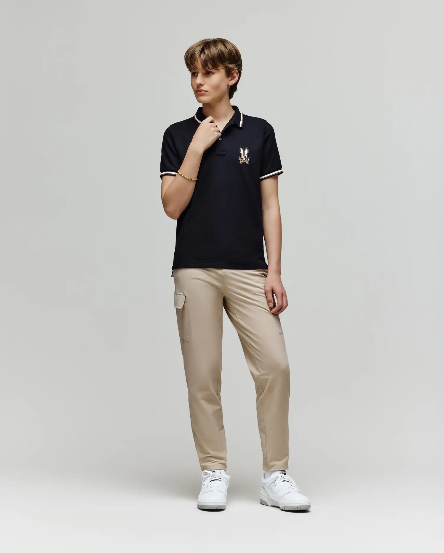A young person stands against a plain background wearing the Psycho Bunny KIDS WADE PIQUE POLO - KT0100032 in black, featuring an embroidered logo, paired with beige cargo pants and white sneakers, looking to the side with one hand on their collar.