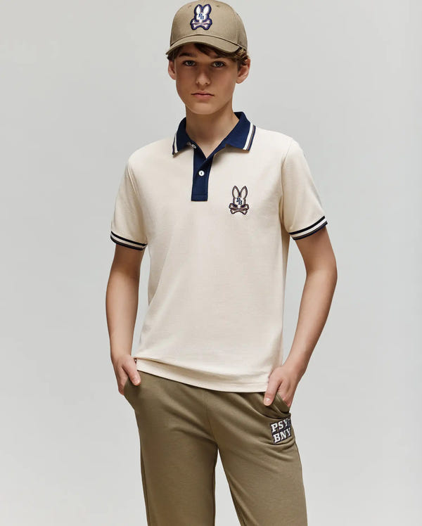 A boy in a Psycho Bunny beige cap with a bunny logo, the KIDS WADE PIQUE POLO - KT0100032 featuring colorblock tipping and an embroidered logo, and khaki joggers with white text, stands against a plain light grey background.
