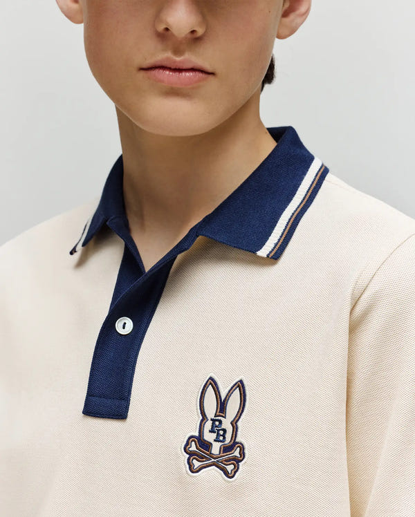 A child wears the Psycho Bunny KIDS WADE PIQUE POLO (KT0100032), a cream Pima cotton shirt with a navy collar, colorblock tipping, and an embroidered bunny skull-and-crossbones logo. The child's face is partially visible from the nose down.