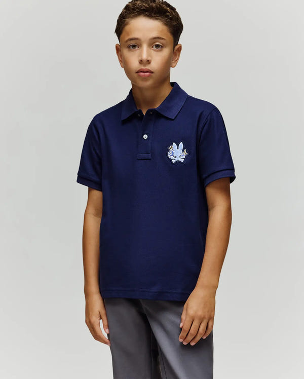 A boy with short curly hair and a neutral expression stands against a plain background, wearing the Psycho Bunny KIDS AUGUSTE PIQUE POLO - KT0100033 in navy blue with a crest on the left chest and gray pants, his left arm relaxed by his side.