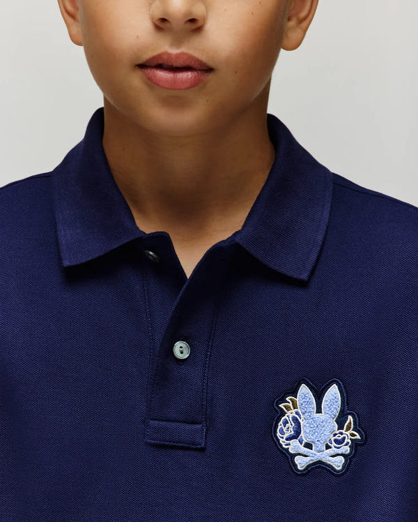 A close-up shows a child wearing the Psycho Bunny KIDS AUGUSTE PIQUE POLO (KT0100033) in navy blue with a light blue bunny and crossbones on the left chest. Only the lower half of the child's face is visible.