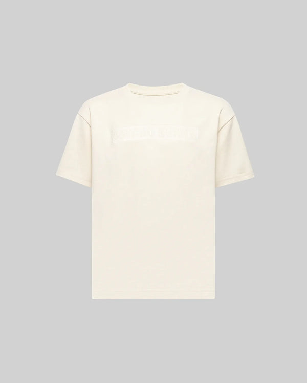 The KIDS ZAYN OVERSIZED GRAPHIC TEE (KT0200001) by Psycho Bunny is a relaxed-fit, off-white, short-sleeve 100% cotton tee featuring a subtle chest patch with indistinct text, shown on a light grey background.