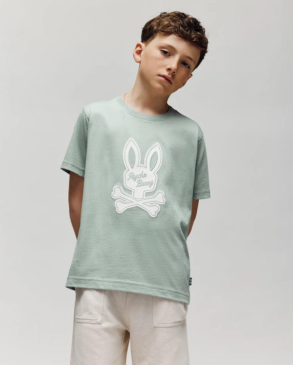 A boy with short brown hair wears the Psycho Bunny KIDS DEREK CENTER GRAPHIC TEE in light green Pima cotton, featuring a large embroidered bunny and crossbones graphic, paired with off-white shorts against a plain light gray background.