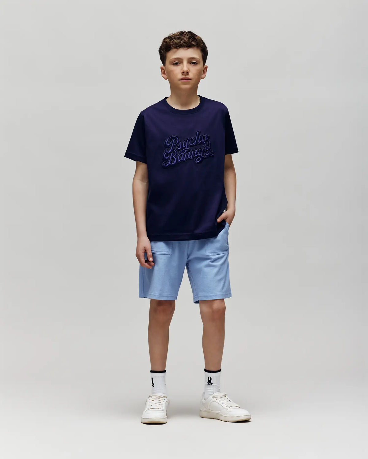 A boy with short brown hair and a neutral expression stands against a plain background, wearing the Psycho Bunny KIDS ELIAS GRAPHIC TEE (KT0200013) in navy, light blue shorts, white socks, and white sneakers, with one hand in his pocket.