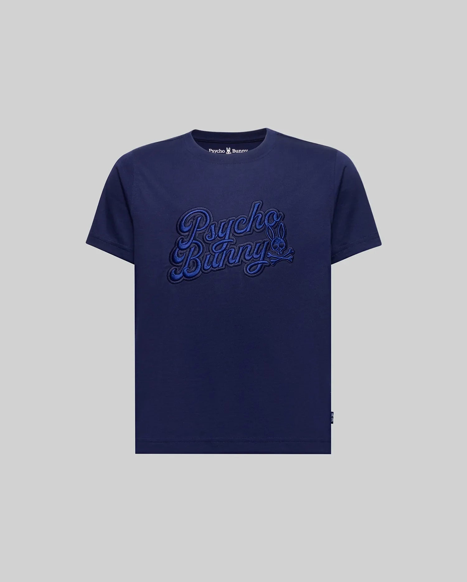 The KIDS ELIAS GRAPHIC TEE (KT0200013) by Psycho Bunny is a navy blue Pima cotton t-shirt featuring bold "Psycho Bunny" script embroidery on the chest in a retro style, displayed on a plain grey background.