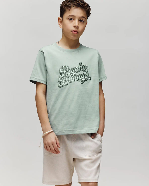 A young boy stands against a plain light background, wearing the Psycho Bunny KIDS ELIAS GRAPHIC TEE (KT0200013) in light green Pima cotton and beige shorts, with one hand in his pocket and a relaxed expression.
