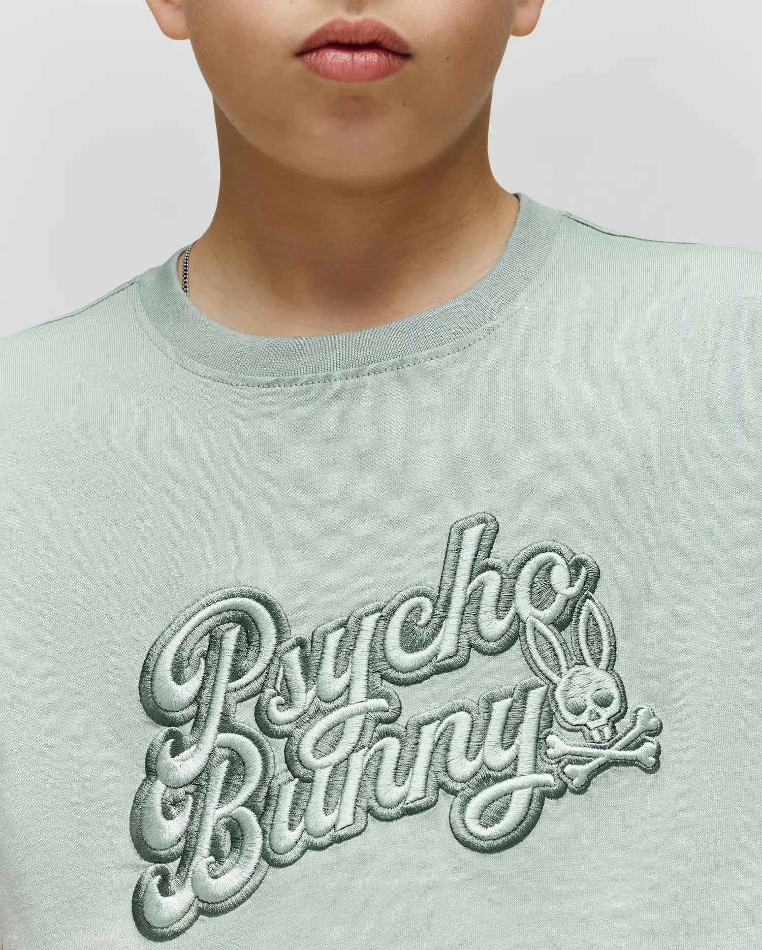A child wears the KIDS ELIAS GRAPHIC TEE by Psycho Bunny, a light green Pima cotton shirt with "Psycho Bunny" lettering and a bold Bunny skull-and-crossbones graphic; only their lower face is visible.