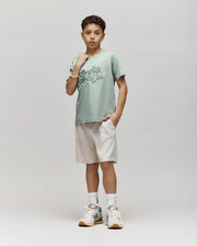 A young boy with short curly hair wears a Psycho Bunny KIDS ELIAS GRAPHIC TEE (KT0200013) in pale green, featuring a bunny graphic, paired with light beige shorts, white socks, and sneakers against a plain background.
