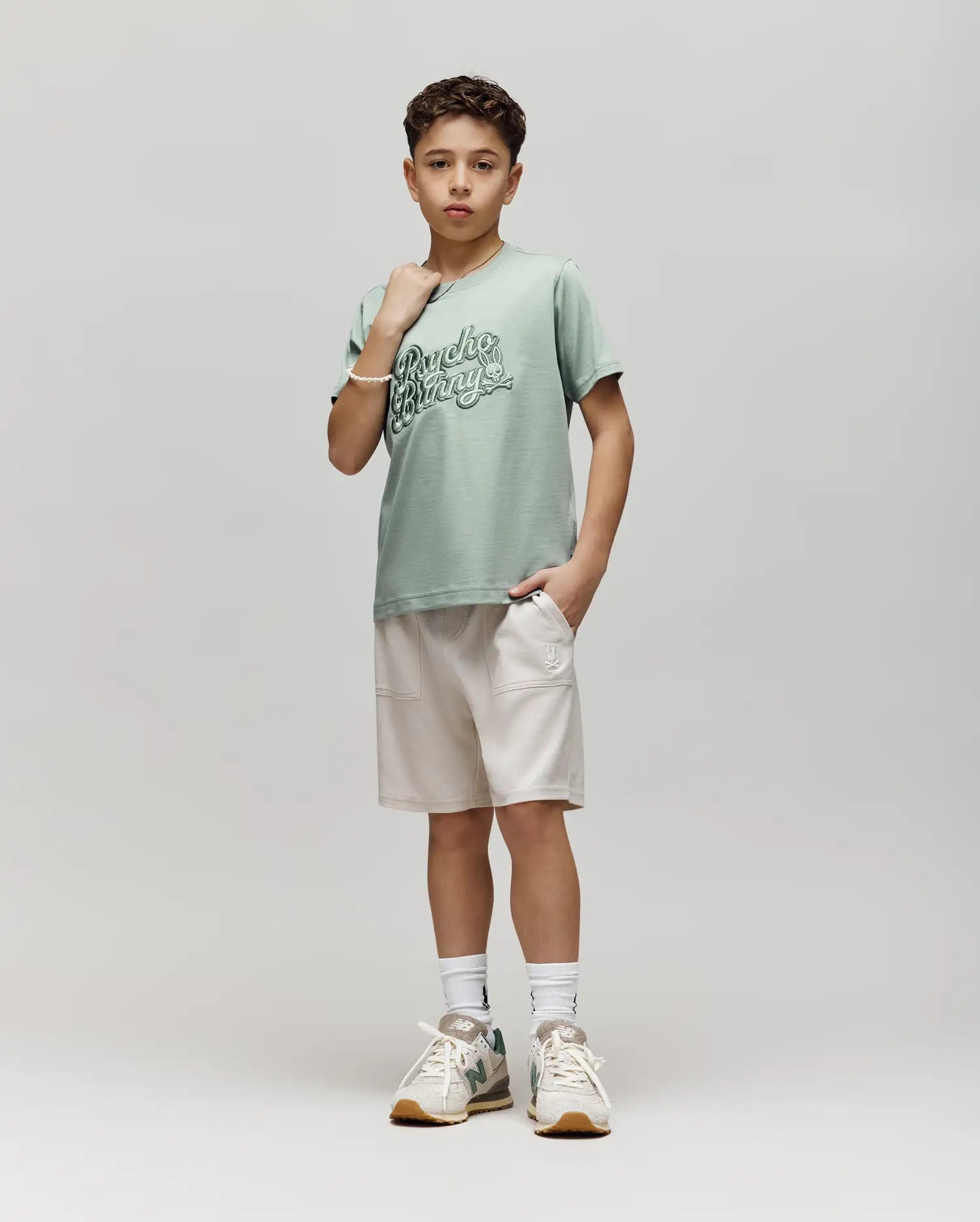 A young boy with short curly hair wears a Psycho Bunny KIDS ELIAS GRAPHIC TEE (KT0200013) in pale green, featuring a bunny graphic, paired with light beige shorts, white socks, and sneakers against a plain background.