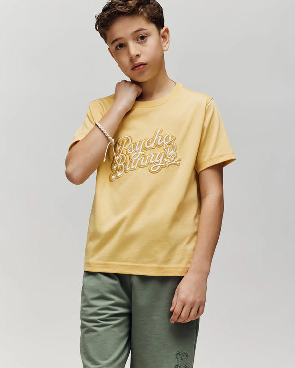 A boy with short brown hair wears the Psycho Bunny KIDS ELIAS GRAPHIC TEE (KT0200013) in yellow, made of soft Pima cotton, with green shorts. He stands against a gray background, relaxed, one hand touching his neck.