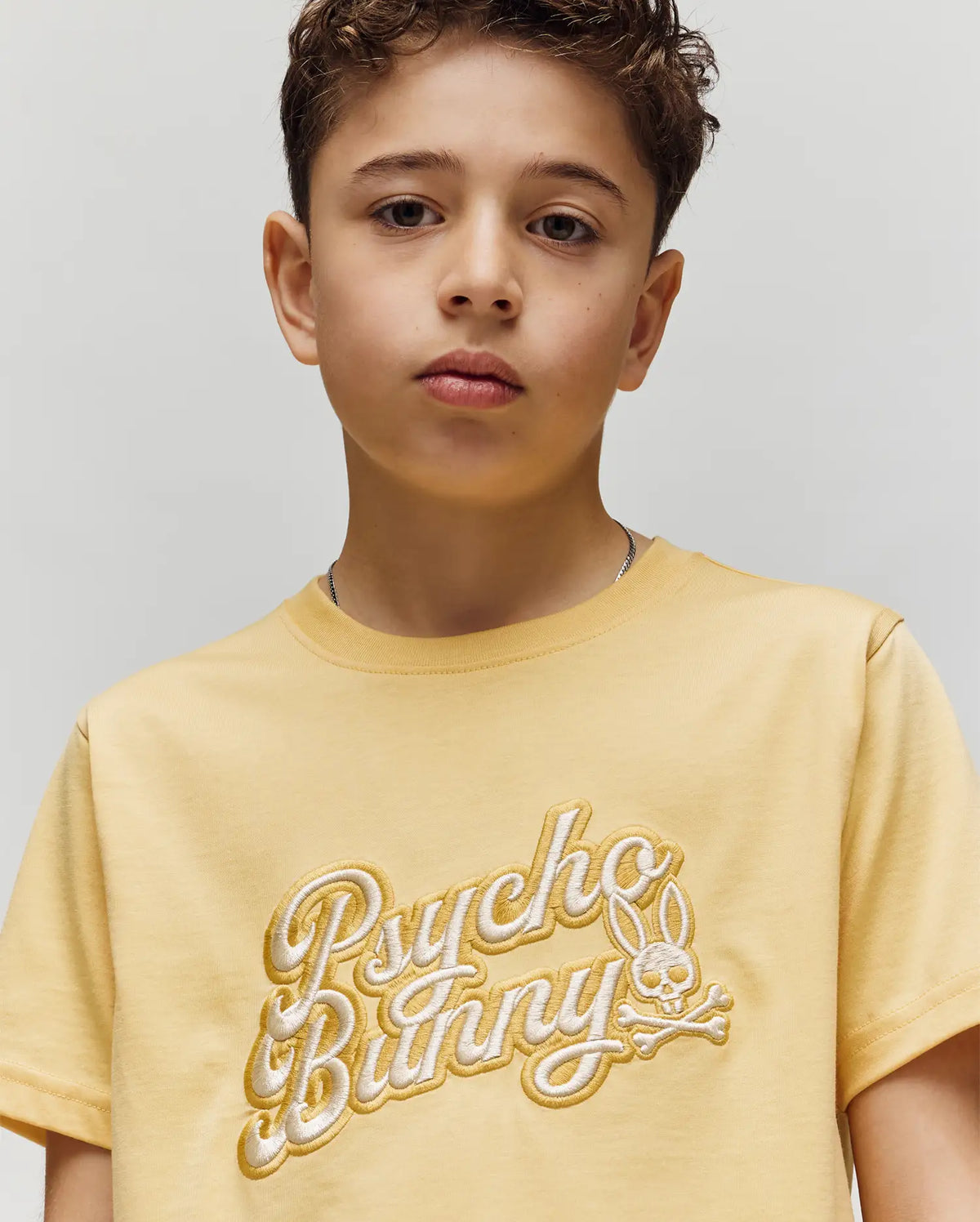 A young boy with short, curly brown hair wears the Psycho Bunny KIDS ELIAS GRAPHIC TEE (KT0200013), a yellow Pima cotton shirt with a Bunny graphic and "Psycho Bunny" logo, standing against a plain light background, looking slightly serious.