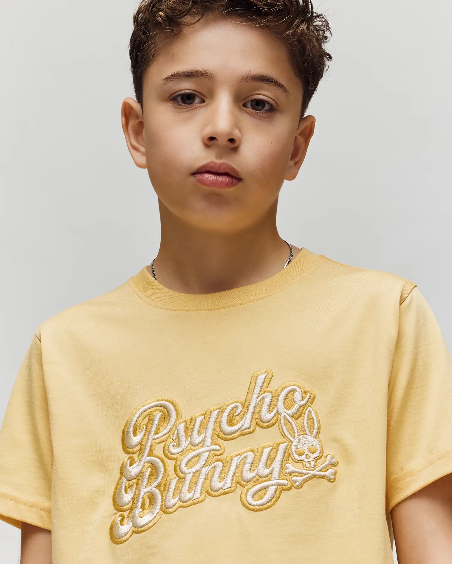 A young boy with short, curly brown hair wears the Psycho Bunny KIDS ELIAS GRAPHIC TEE (KT0200013), a yellow Pima cotton shirt with a Bunny graphic and "Psycho Bunny" logo, standing against a plain light background, looking slightly serious.