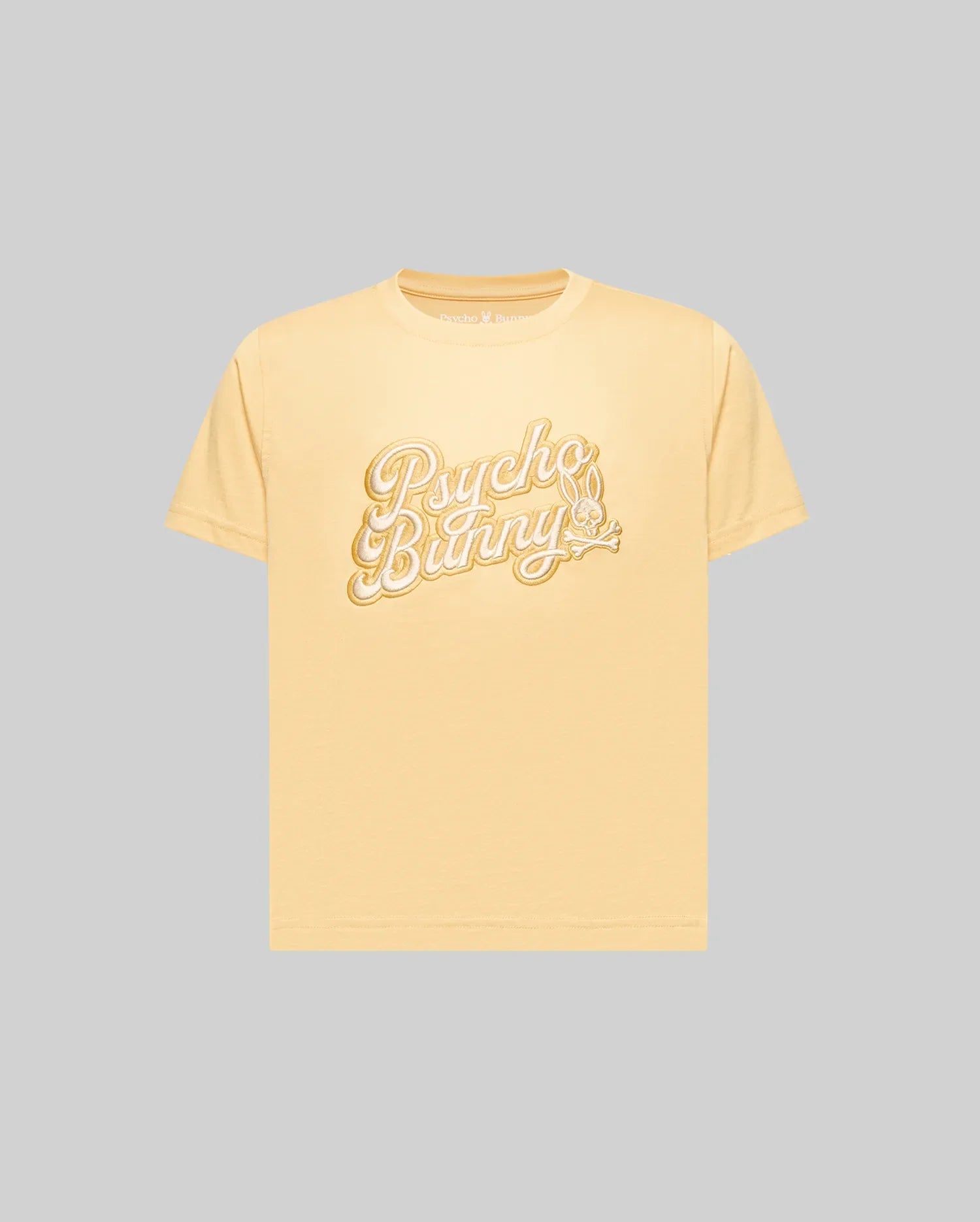 The KIDS ELIAS GRAPHIC TEE (KT0200013) by Psycho Bunny is a light yellow, retro-style short-sleeve Pima cotton t-shirt featuring "Psycho Bunny" in large white script and a bunny with a carrot, shown on a plain gray background.
