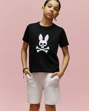 A young person stands against a pink background wearing the Psycho Bunny KIDS ARCHIVE BUNNY GRAPHIC TEE (KT0200019), a black Pima cotton T-shirt with an embroidered Bunny skull and crossbones, rib knit crewneck, paired with light shorts.