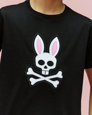 A person wears the Psycho Bunny KIDS ARCHIVE BUNNY GRAPHIC TEE (KT0200019), a black Pima cotton t-shirt with a large skull featuring pink and white bunny ears and crossbones, rib knit crewneck, against a light pink background.