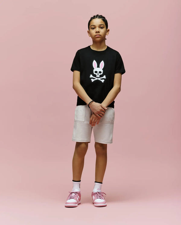 A young person stands against a pink background, wearing the Psycho Bunny KIDS ARCHIVE BUNNY GRAPHIC TEE (KT0200019), black Pima cotton with an embroidered bunny skull, paired with light shorts, white socks, and pink sneakers.