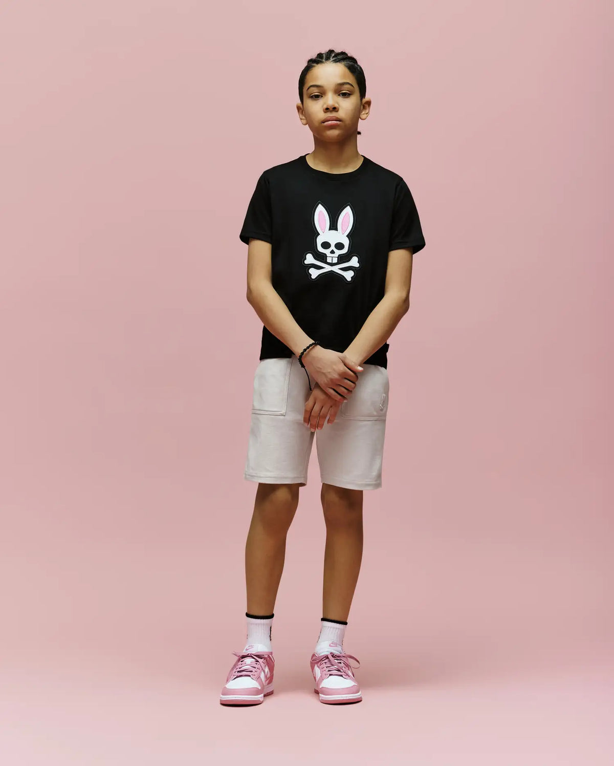 A young person stands against a pink background, wearing the Psycho Bunny KIDS ARCHIVE BUNNY GRAPHIC TEE (KT0200019), black Pima cotton with an embroidered bunny skull, paired with light shorts, white socks, and pink sneakers.