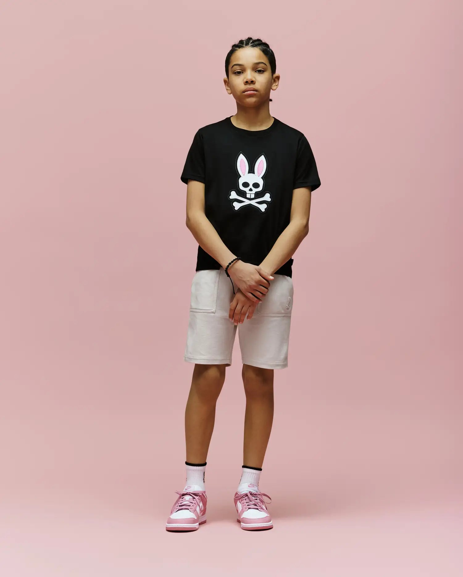 A young person stands against a pink background, wearing the Psycho Bunny KIDS ARCHIVE BUNNY GRAPHIC TEE (KT0200019), black Pima cotton with an embroidered bunny skull, paired with light shorts, white socks, and pink sneakers.