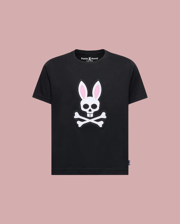 The Psycho Bunny KIDS ARCHIVE BUNNY GRAPHIC TEE (KT0200019) is a black Pima cotton t-shirt with a skull and crossbones bunny graphic with pink ears, shown on a solid light pink background.