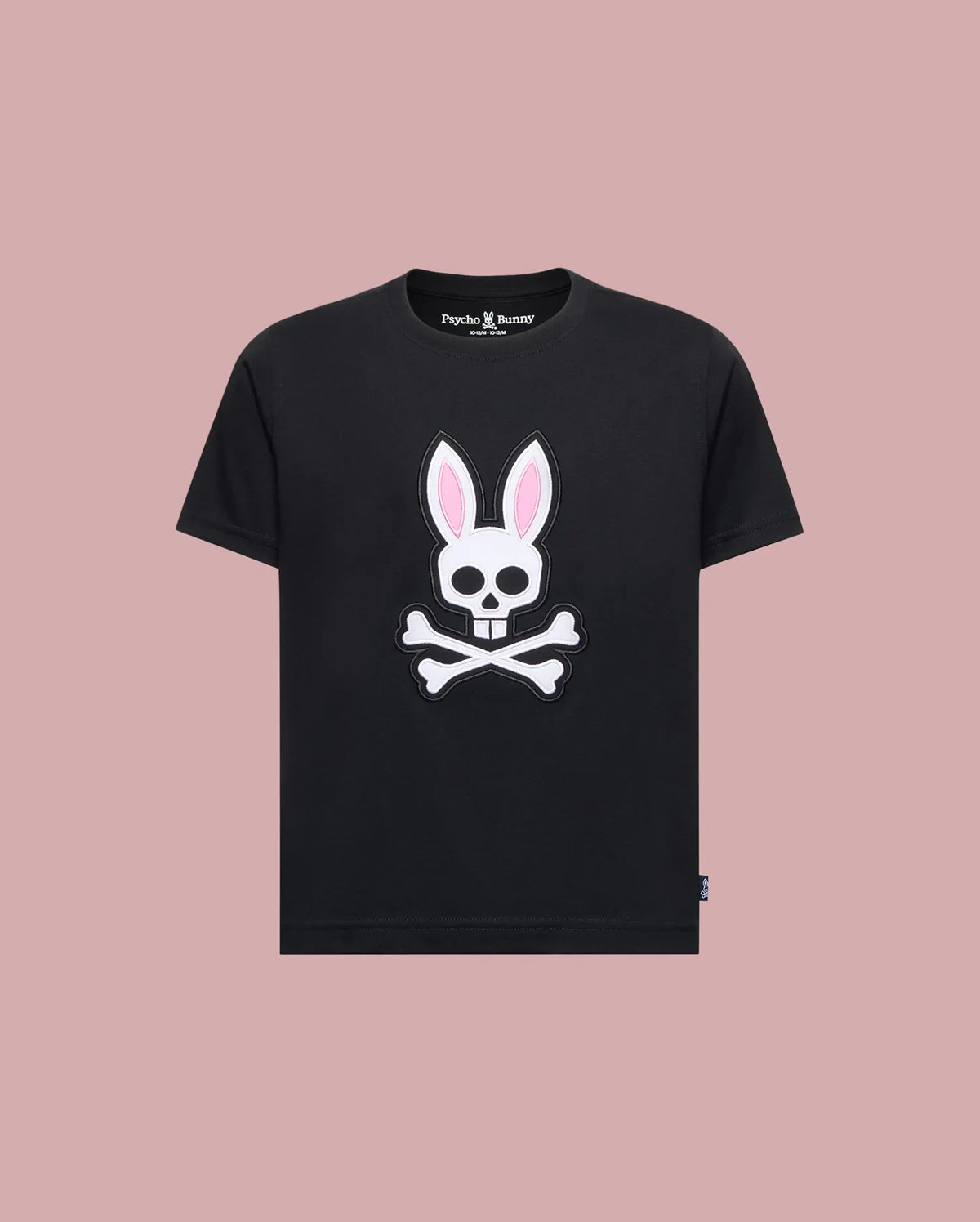The Psycho Bunny KIDS ARCHIVE BUNNY GRAPHIC TEE (KT0200019) is a black Pima cotton t-shirt with a skull and crossbones bunny graphic with pink ears, shown on a solid light pink background.