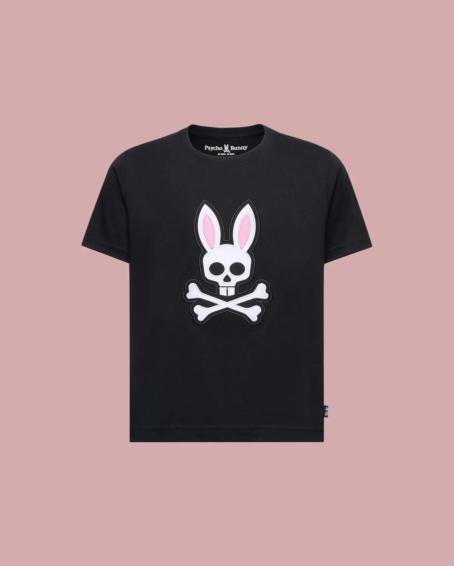 The Psycho Bunny KIDS ARCHIVE BUNNY GRAPHIC TEE (KT0200019) is a black Pima cotton t-shirt with a skull and crossbones bunny graphic with pink ears, shown on a solid light pink background.
