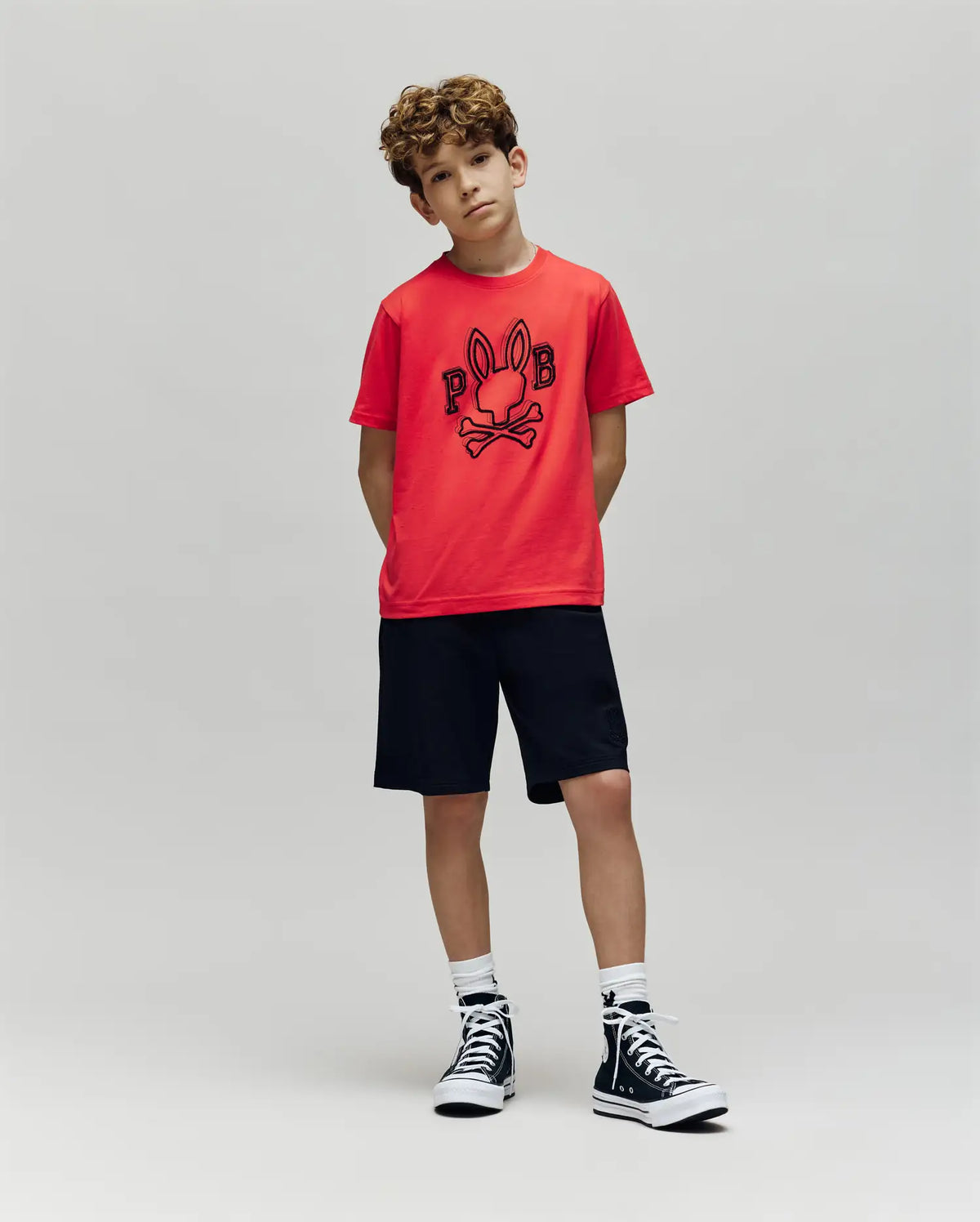 A young boy with curly hair stands against a gray background, wearing the Psycho Bunny KIDS BALES GRAPHIC TEE (KT0200021) in red with a Big Bunny print, black shorts, white socks, and black high-tops—ideal for year-round style.