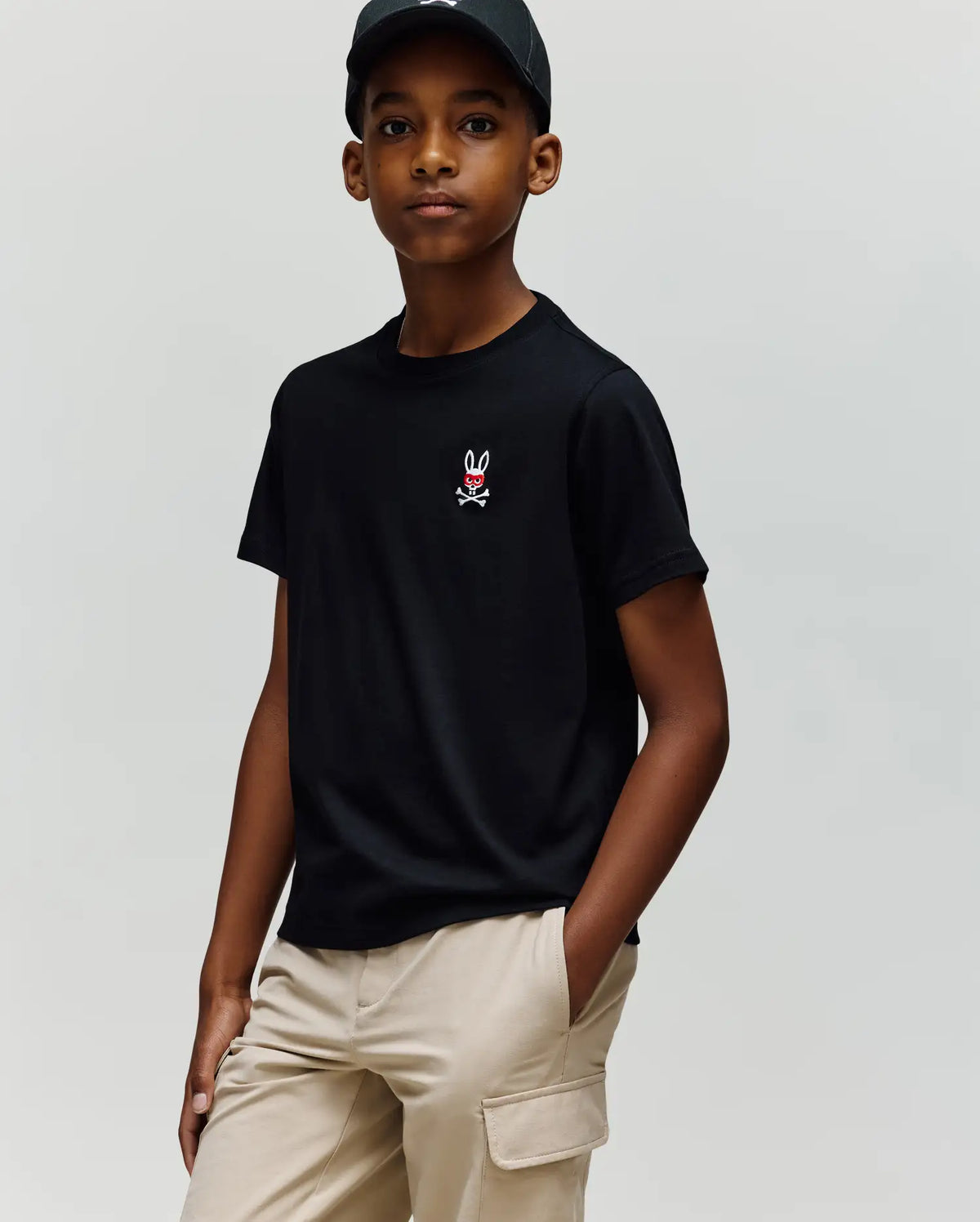 A young person wearing the Psycho Bunny KIDS MISCHIEF BUNNY TEE (KT0200023) in black Pima cotton with an embroidered Mischief Bunny logo and beige cargo pants stands with one hand in their pocket against a plain background.