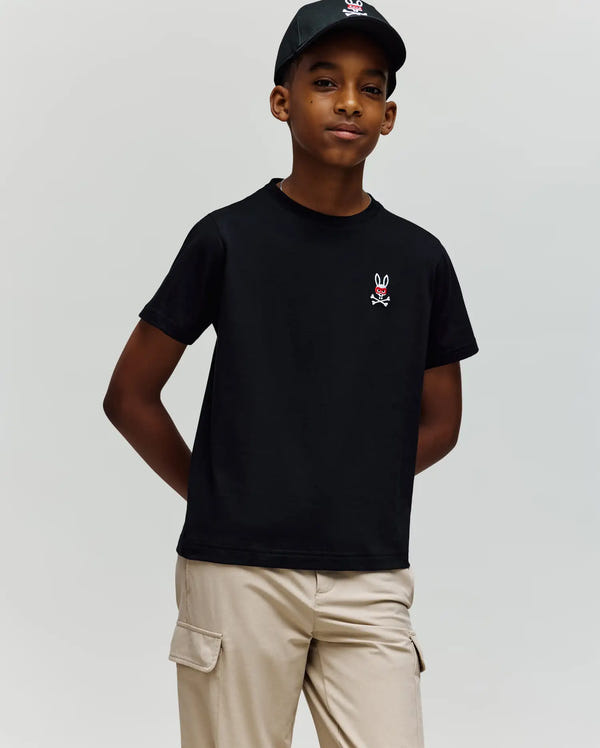A boy stands against a plain background with hands in his pockets, wearing the Psycho Bunny KIDS MISCHIEF BUNNY TEE (KT0200023) in black Pima cotton featuring an embroidered Mischief Bunny and crossbones logo, paired with light beige cargo pants.