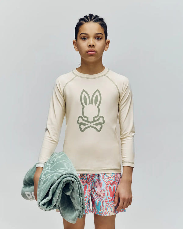 A child with braided hair stands against a plain background, wearing Psycho Bunny’s KIDS LONG SLEEVE RASHGUARD - KT0200027 in beige with a Bunny and crossbones logo, patterned swim trunks, and holding a folded green towel. The rash guard offers UPF 50+ protection.