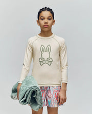 A child with braided hair stands against a plain background, wearing Psycho Bunny’s KIDS LONG SLEEVE RASHGUARD - KT0200027 in beige with a Bunny and crossbones logo, patterned swim trunks, and holding a folded green towel. The rash guard offers UPF 50+ protection.