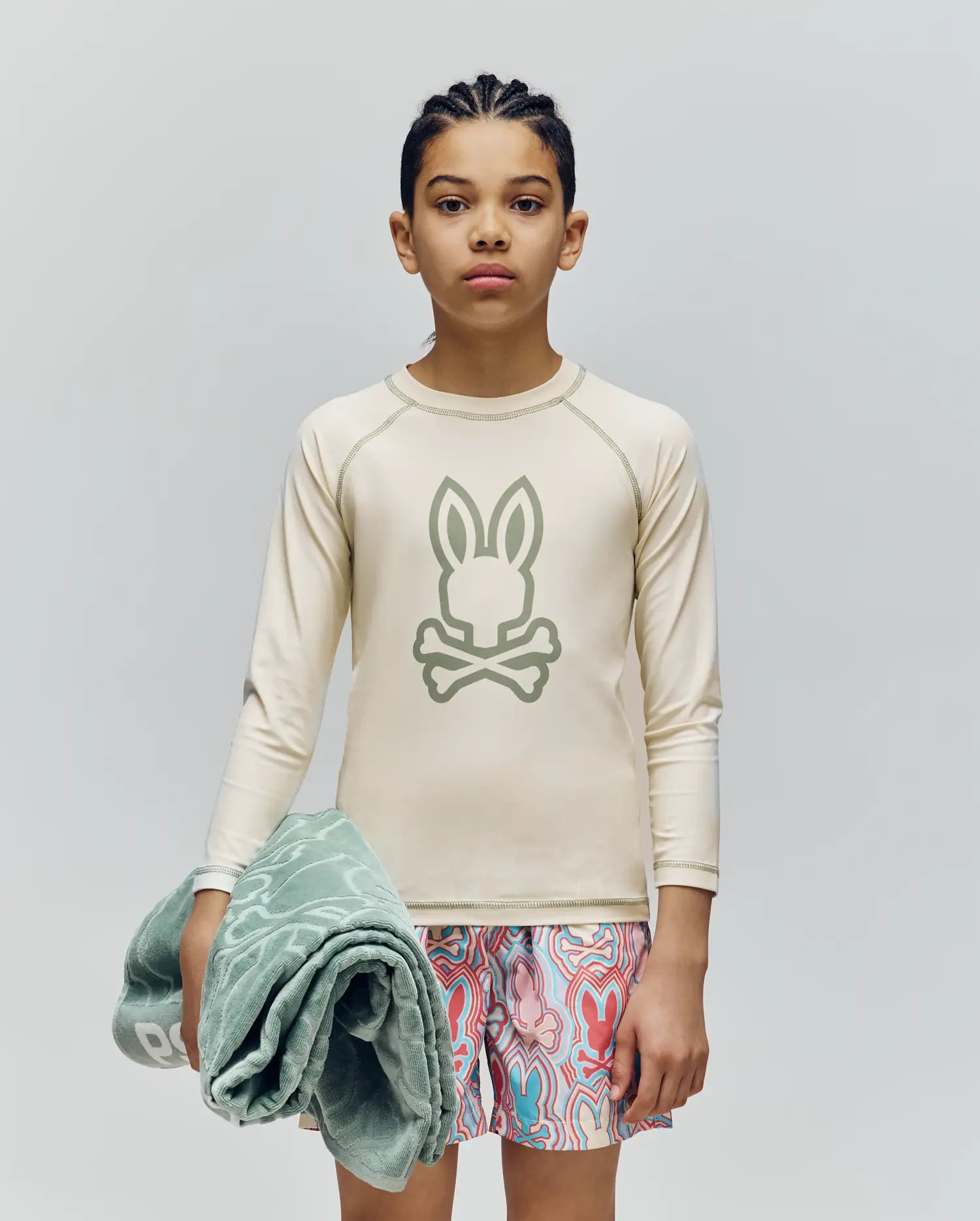 A child with braided hair stands against a plain background, wearing Psycho Bunny’s KIDS LONG SLEEVE RASHGUARD - KT0200027 in beige with a Bunny and crossbones logo, patterned swim trunks, and holding a folded green towel. The rash guard offers UPF 50+ protection.