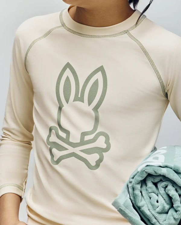 A child wears the Psycho Bunny KIDS LONG SLEEVE RASHGUARD (KT0200027) in beige, showcasing a bold bunny and crossbones print on the front while holding a folded light green towel; only their torso and lower face are visible.