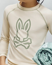 A child wears the Psycho Bunny KIDS LONG SLEEVE RASHGUARD (KT0200027) in beige, showcasing a bold bunny and crossbones print on the front while holding a folded light green towel; only their torso and lower face are visible.