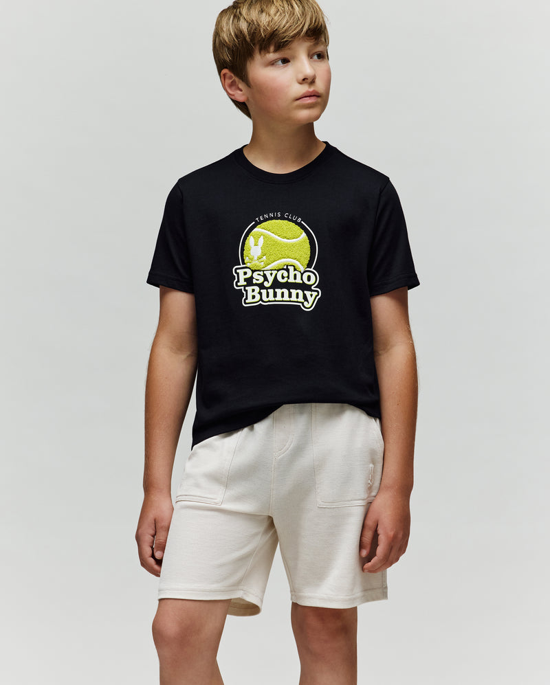 A young boy in white shorts wears the Psycho Bunny KIDS PETE TENNIS GRAPHIC TEE (KT0200038), a classic fit Pima cotton shirt featuring a tennis ball logo, standing against a plain light background and looking to the side.