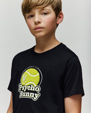 A young boy with light brown hair wears the Psycho Bunny KIDS PETE TENNIS GRAPHIC TEE (KT0200038), featuring a tennis ball graphic and "Psycho Bunny Tennis Club" text, against a plain light background.