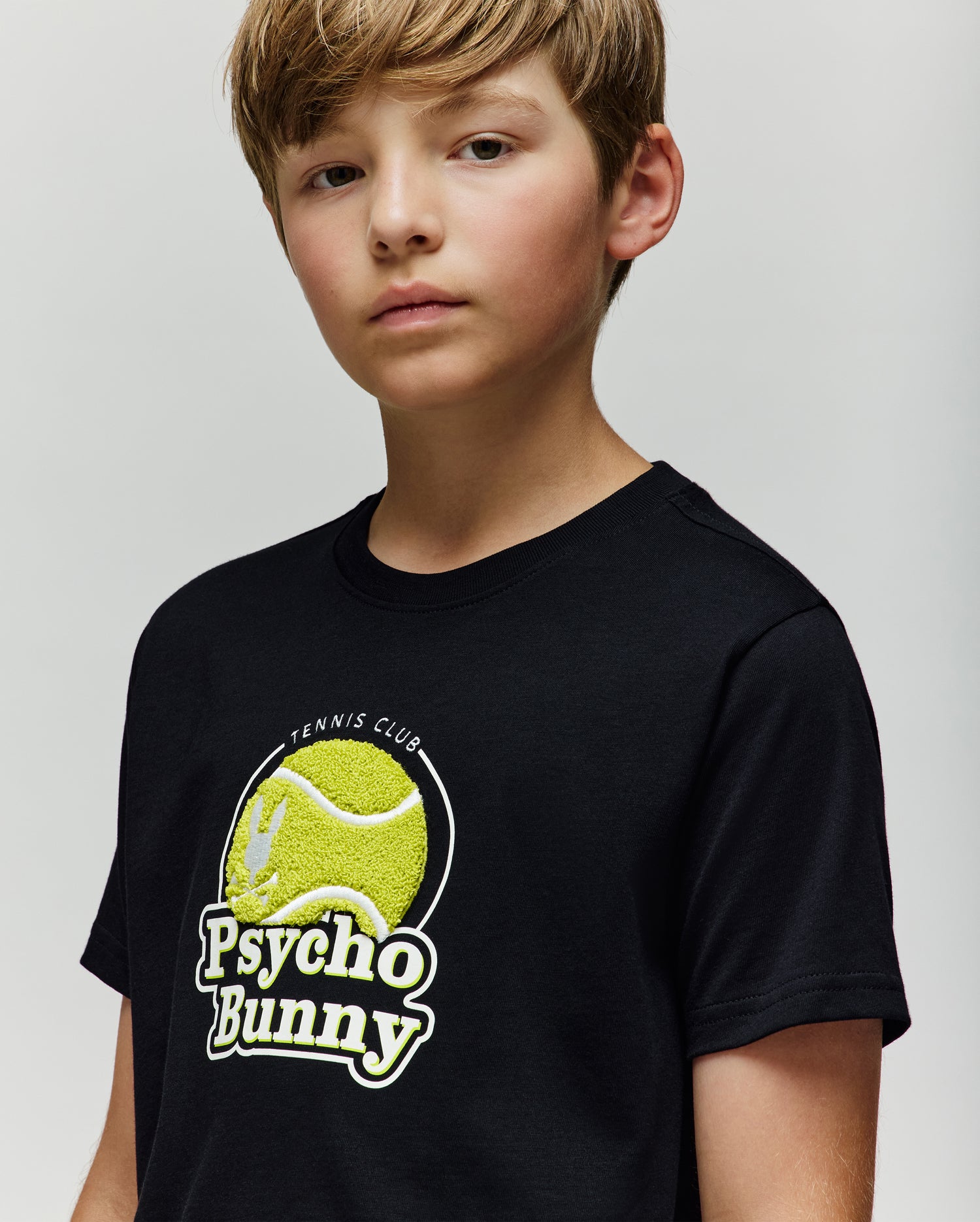 A young boy with light brown hair wears the Psycho Bunny KIDS PETE TENNIS GRAPHIC TEE (KT0200038), featuring a tennis ball graphic and "Psycho Bunny Tennis Club" text, against a plain light background.
