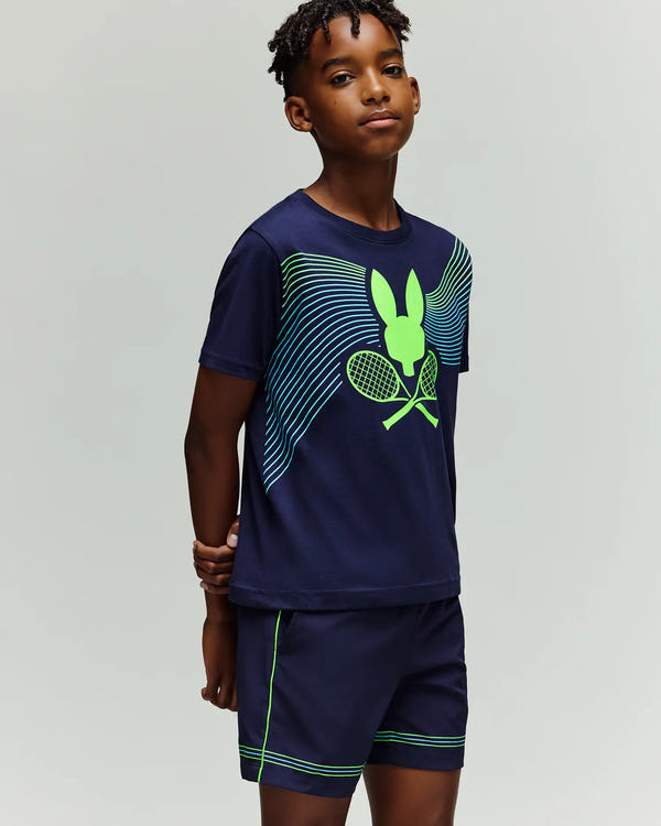 A young boy stands against a light grey background, wearing Psycho Bunny's RETAIL BALL CREW YOUTH TEE (KT0200041) in navy blue with a neon green bunny and crossed tennis rackets, paired with matching navy shorts with green accents.