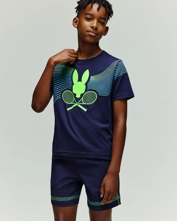 A boy stands against a light gray background wearing the Psycho Bunny RETAIL BALL CREW YOUTH TEE (EN) - KT0200041, a navy blue T-shirt and shorts with neon green accents and a graphic of a rabbit head and crossed tennis rackets.