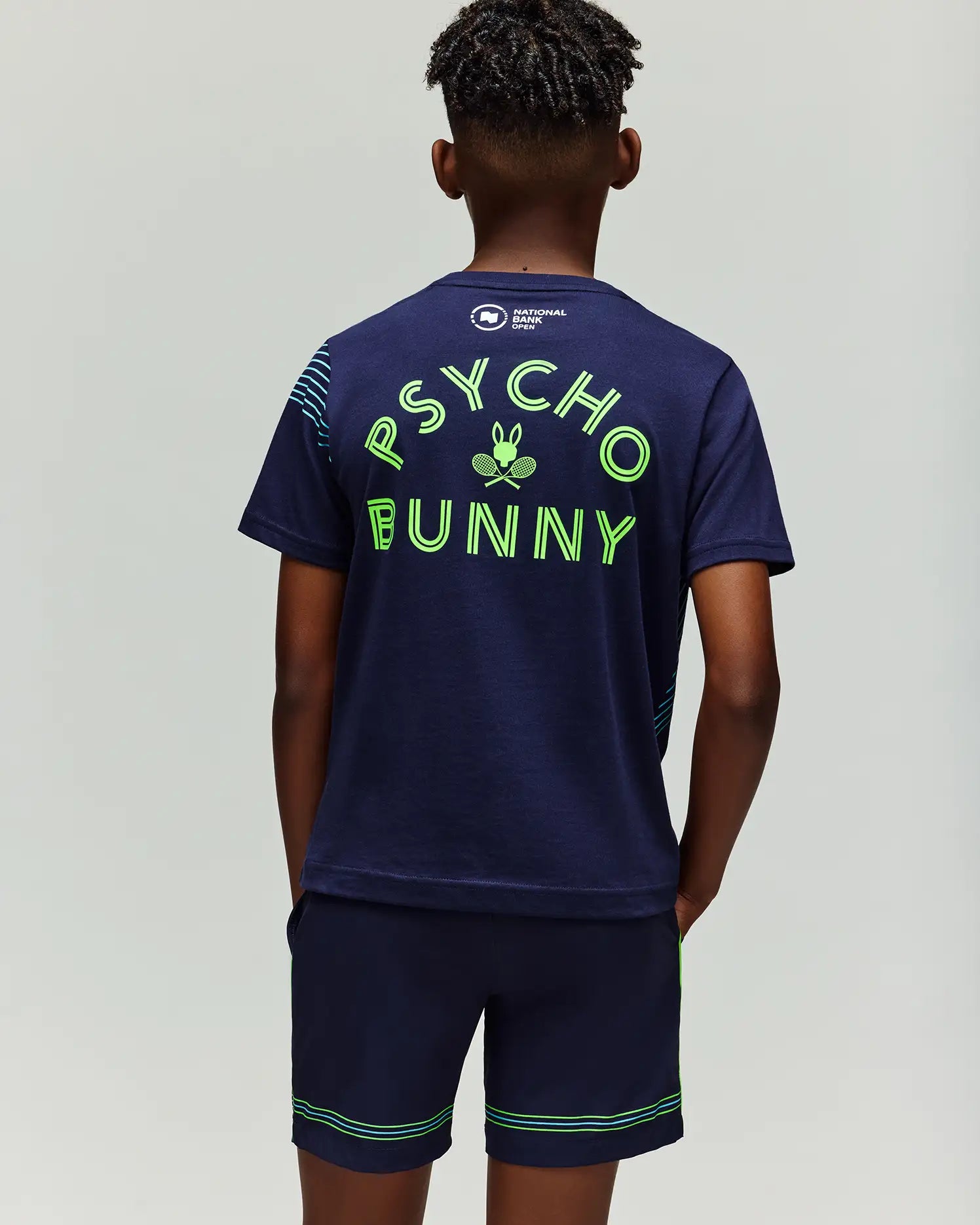 A person stands facing away, wearing the Psycho Bunny RETAIL BALL CREW YOUTH TEE in navy blue. The t-shirt features a large neon green "PSYCHO BUNNY" and bunny logo printed on the back. Neon-trimmed shorts complete the look.