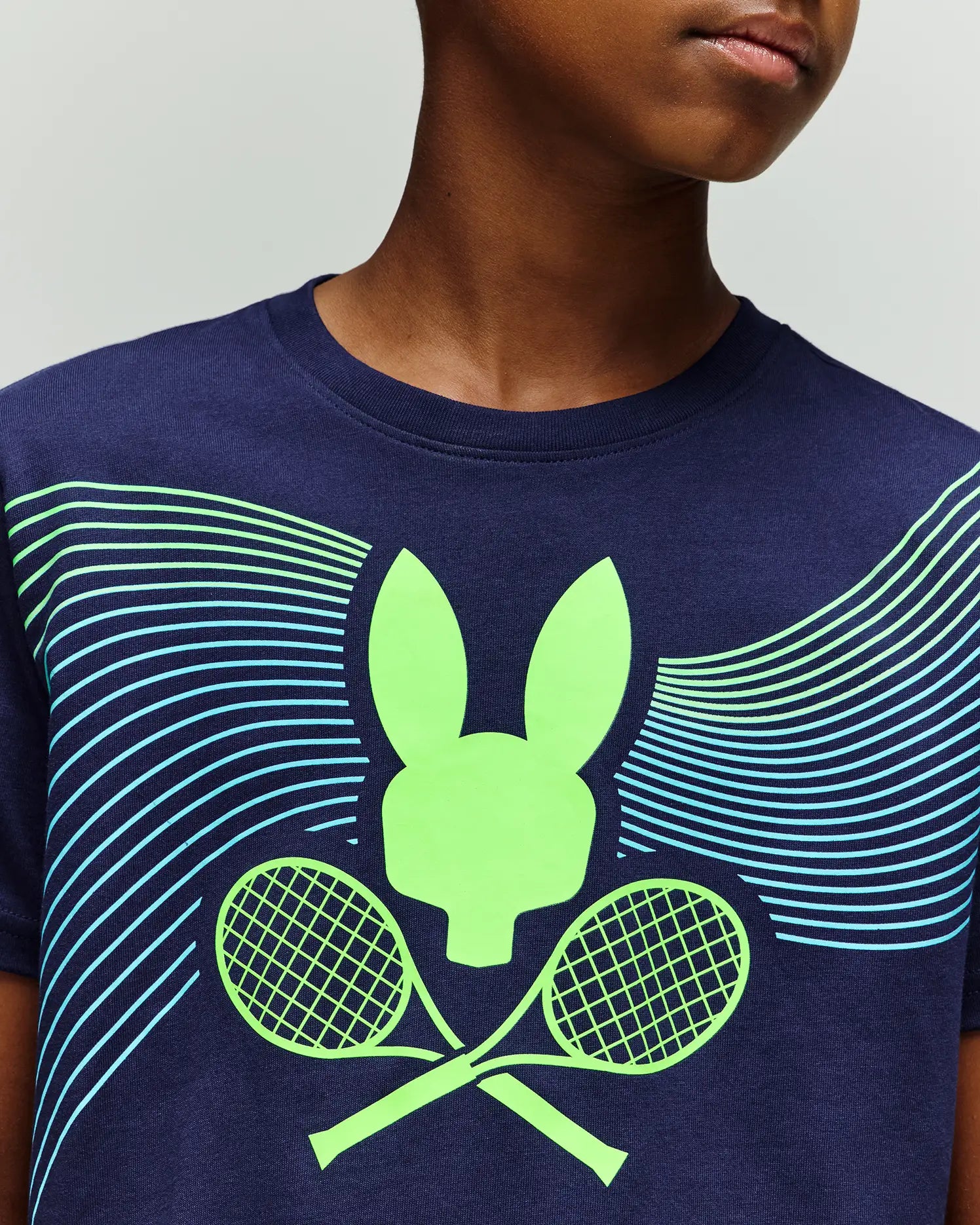 A child wears the Psycho Bunny RETAIL BALL CREW YOUTH TEE (FR) - KT0200042, a navy blue shirt featuring a neon green bunny head and crossed tennis rackets graphic with curved blue and green lines.