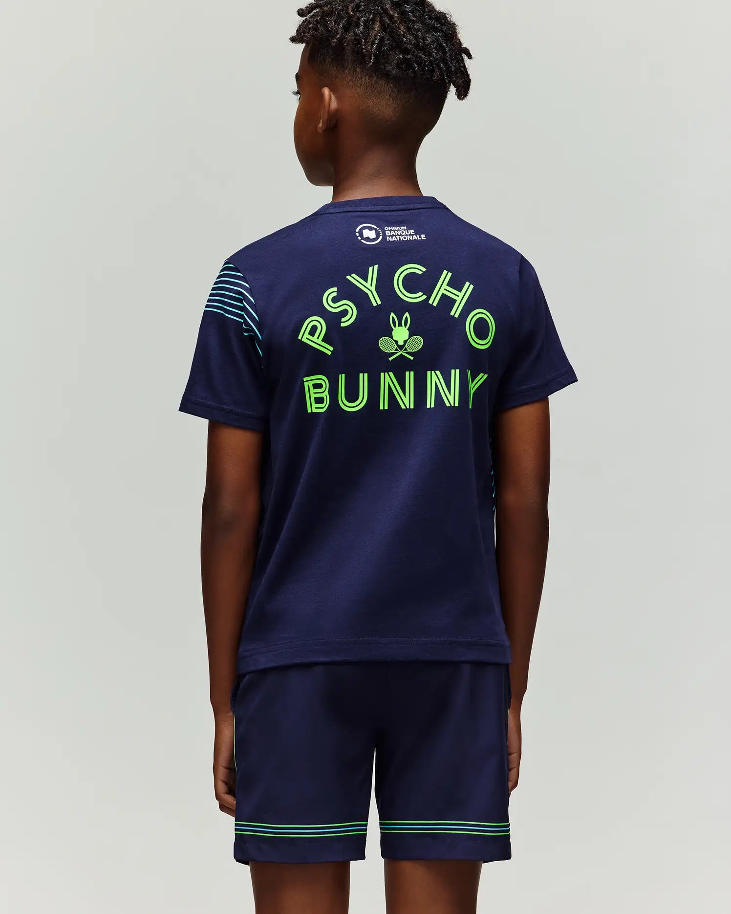 A boy faces away wearing the Psycho Bunny RETAIL BALL CREW YOUTH TEE (FR) - KT0200042, a navy blue t-shirt with "PSYCHO BUNNY" in bright green letters and a bunny with crossed racquets graphic below.