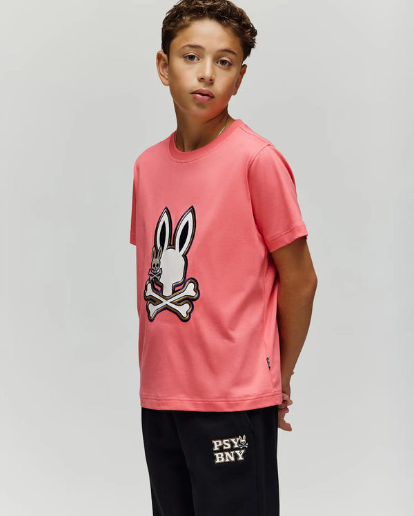 A boy with curly hair wears the Psycho Bunny KIDS WADE GRAPHIC TEE (KT0200054), a pink 100% cotton shirt featuring a cartoon bunny and crossbones, paired with black "PSY BNY" shorts, standing against a plain light background.