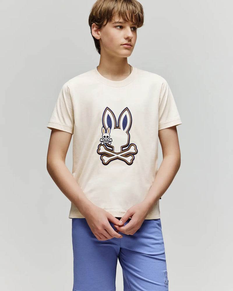 A teenage boy wears the Psycho Bunny KIDS WADE GRAPHIC TEE - KT0200054, a cream-colored shirt with an embroidered Bunny and crossbones design, paired with light blue shorts, standing against a plain light gray background.