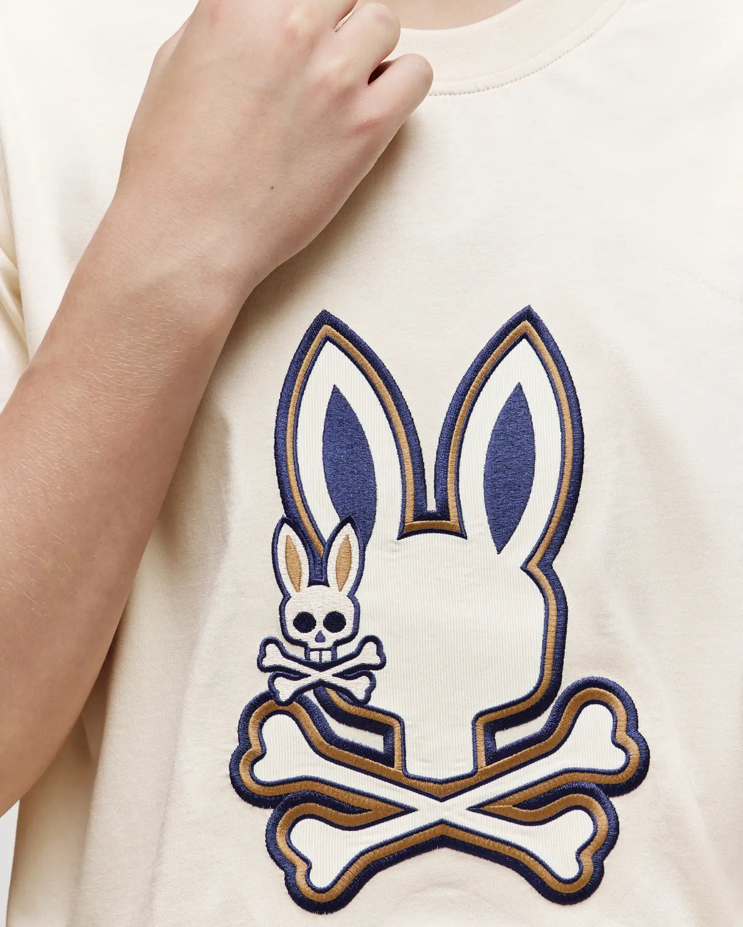 Someone wearing the Psycho Bunny KIDS WADE GRAPHIC TEE—KT0200054, a cream-colored 100% cotton shirt with a large embroidered Bunny head above crossed bones and a smaller bunny skull and crossbones on the left side.
