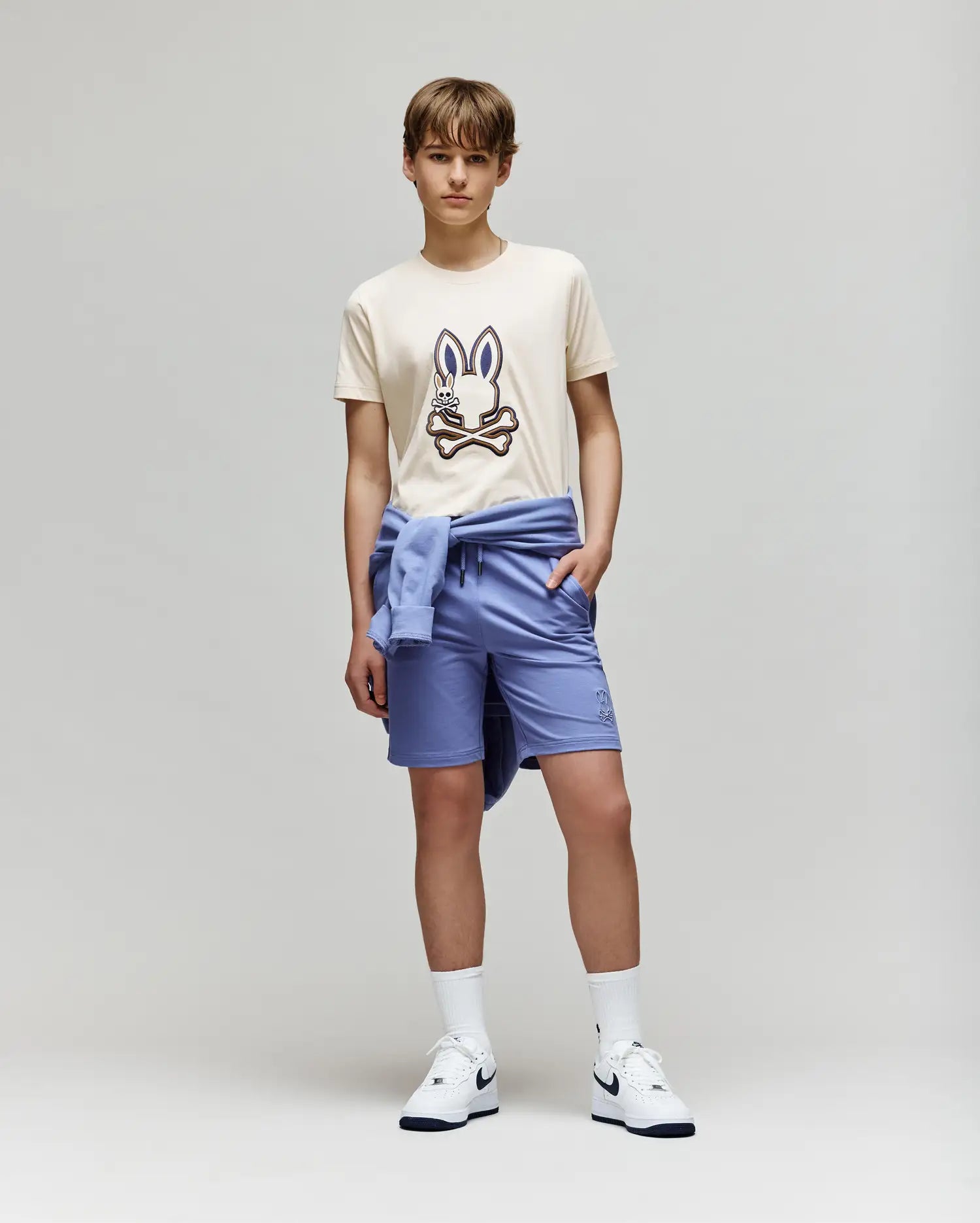 A teenage boy stands against a plain background wearing the Psycho Bunny KIDS WADE GRAPHIC TEE - KT0200054 in beige with an embroidered bunny, paired with light blue shorts, a matching sweatshirt tied around his waist, white socks, and Nike sneakers.
