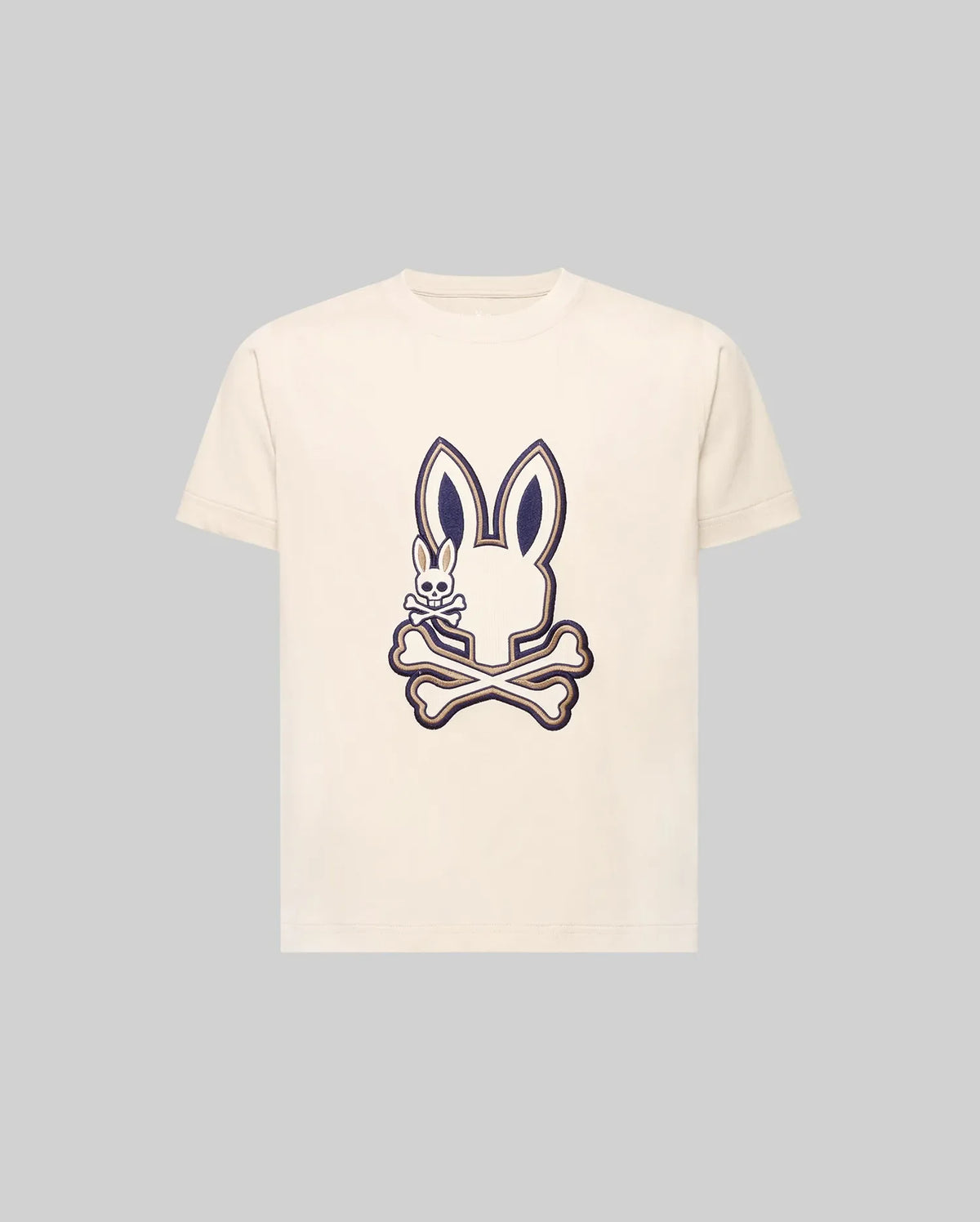 The KIDS WADE GRAPHIC TEE - KT0200054 by Psycho Bunny is a cream-colored 100% cotton tee with a large bunny head and crossbones graphic, plus a small smiling embroidered Bunny on the chest. The background is light gray.