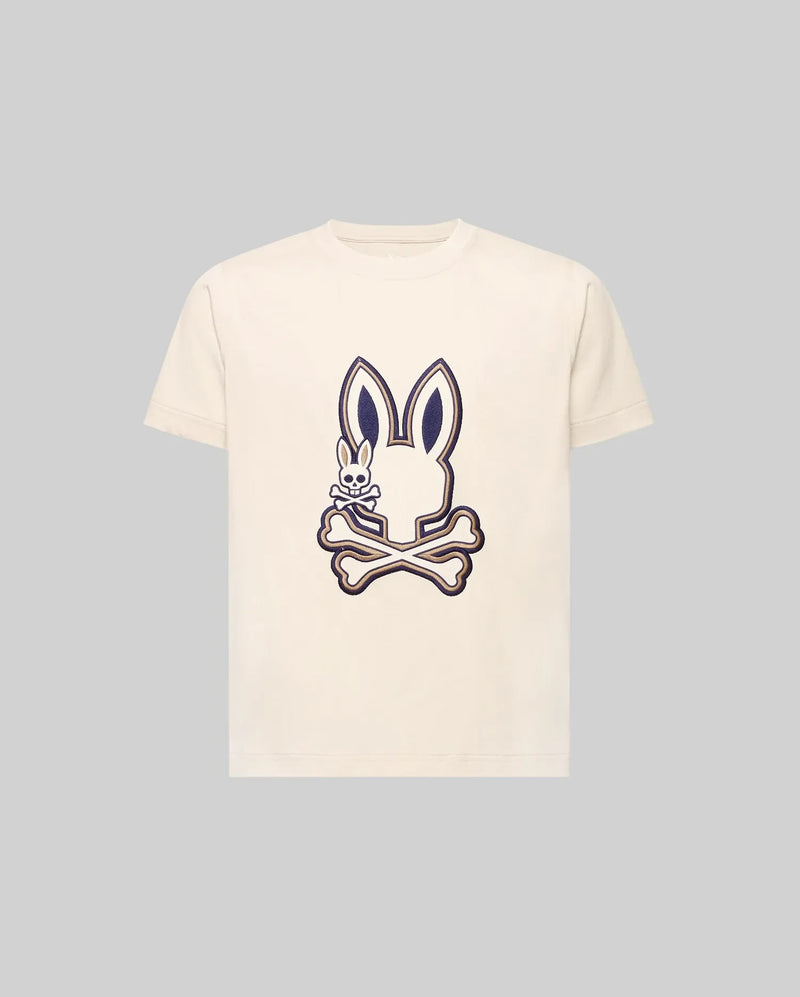 The KIDS WADE GRAPHIC TEE - KT0200054 by Psycho Bunny is a cream-colored 100% cotton tee with a large bunny head and crossbones graphic, plus a small smiling embroidered Bunny on the chest. The background is light gray.